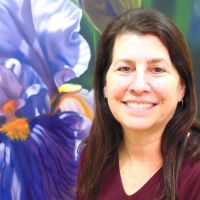 Woman with long brown hair and red shirt stands in front of a botanical illustration of a purple flower