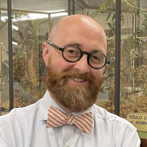 A man with glasses, a beard, and bowtie stands in front of a glass display case